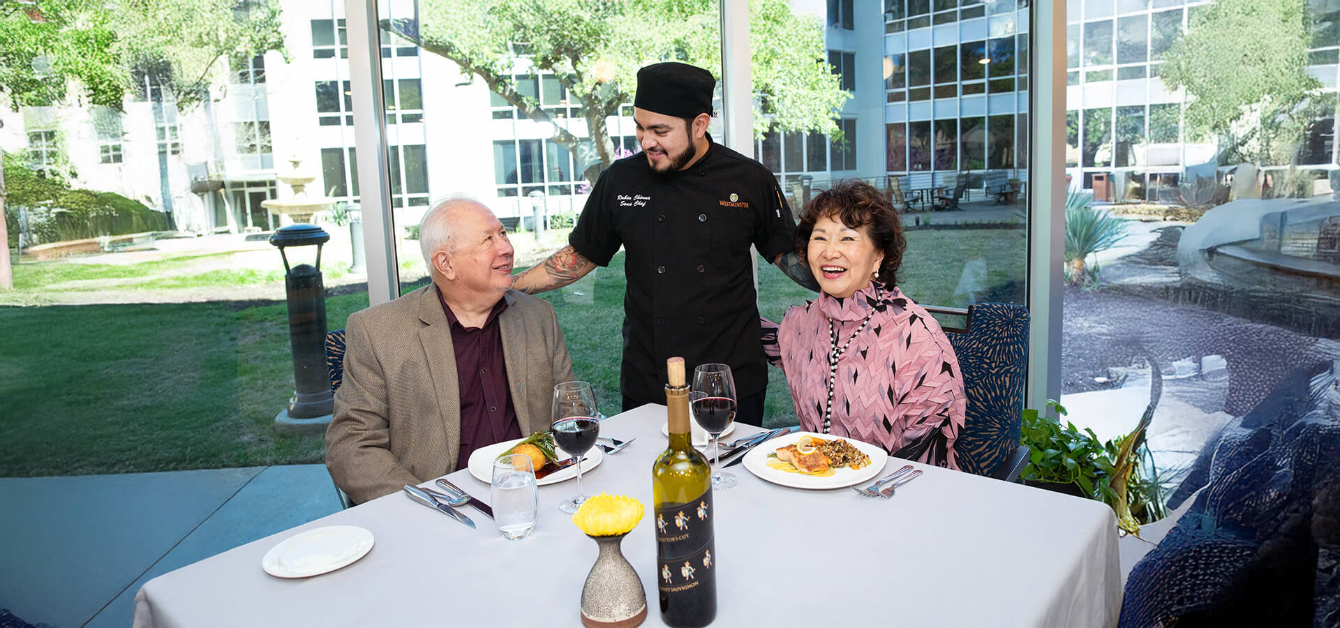 Couple dines together at a table while smiling chef looks on