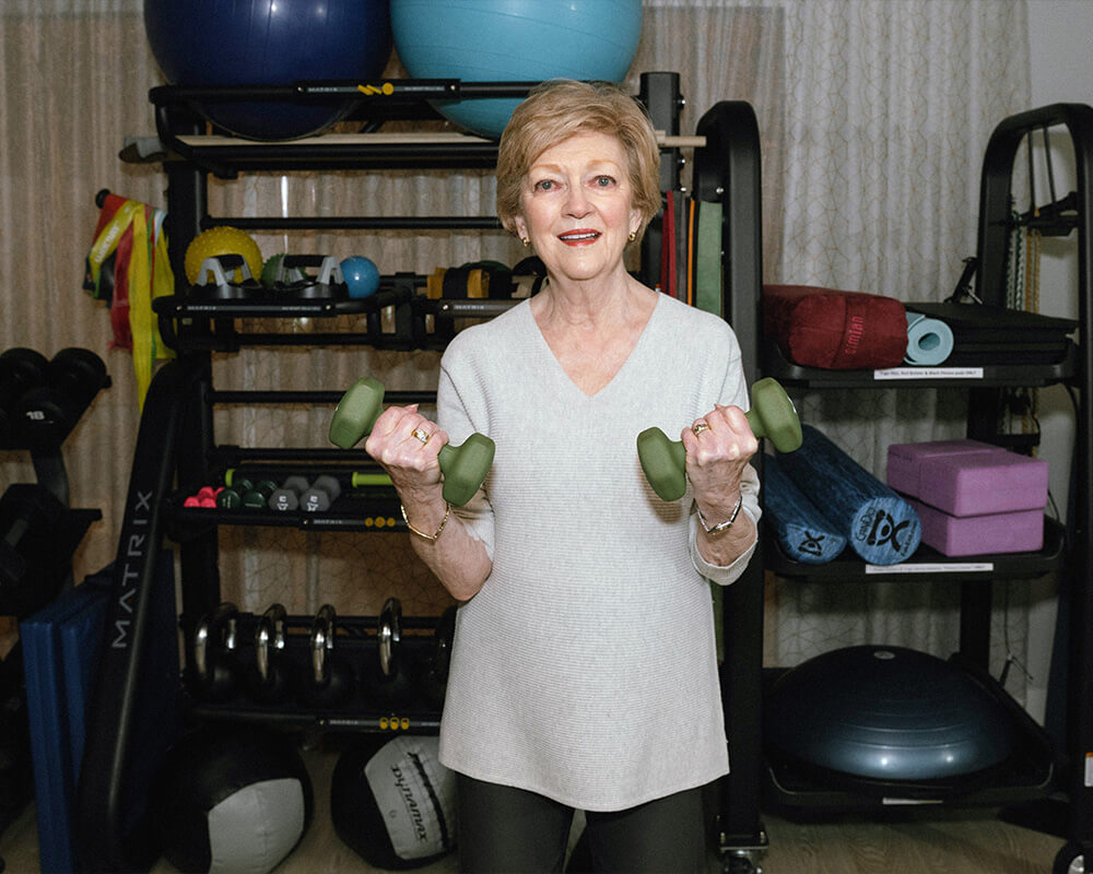 Woman lifting weights in a fitness room with exercise equipment in the background.