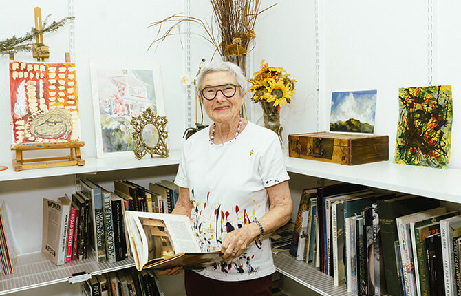 Elderly woman in a library reading a book surrounded by art and books.
