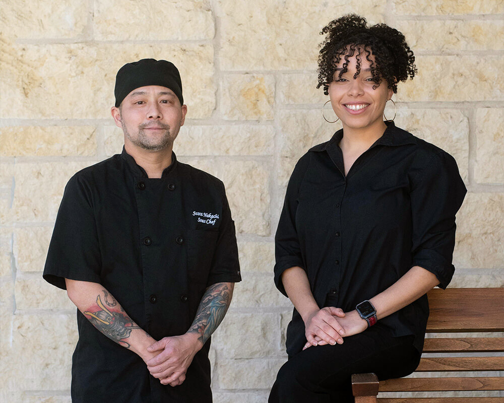 Two staff members in black uniforms, smiling in front of a stone wall.