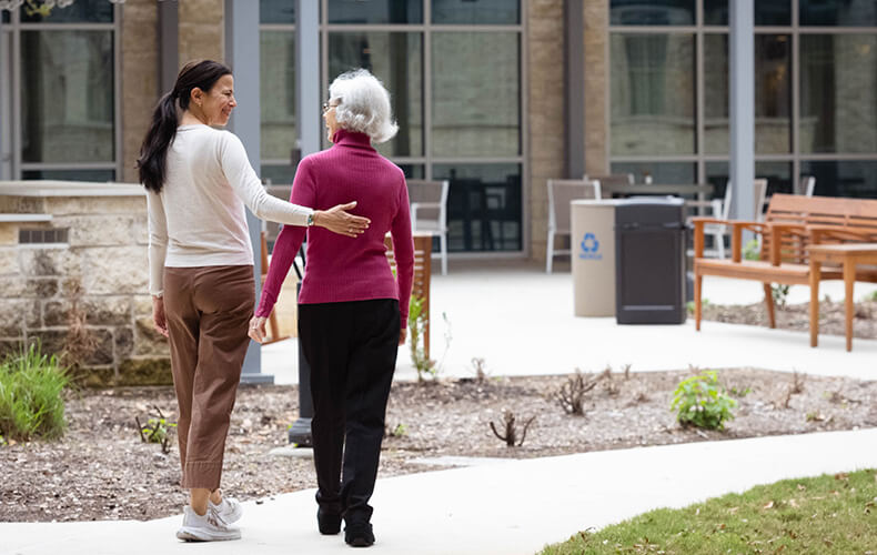 Two women walking together in a courtyard of a senior living facility.