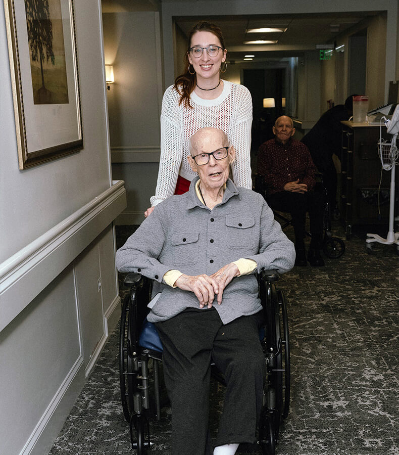 Caregiver standing behind resident in wheelchair within a senior living unit hallway.