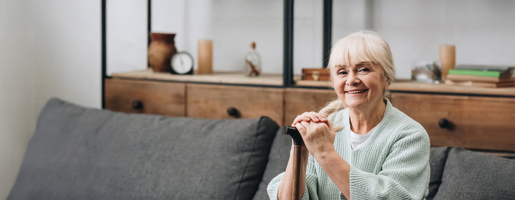 Smiling senior woman with a cane seated in a cozy unit interior.