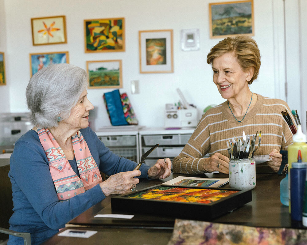 Two senior women engaged in art activities in a well-lit studio filled with paintings.