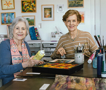 Two women painting at a table in a brightly decorated art room.