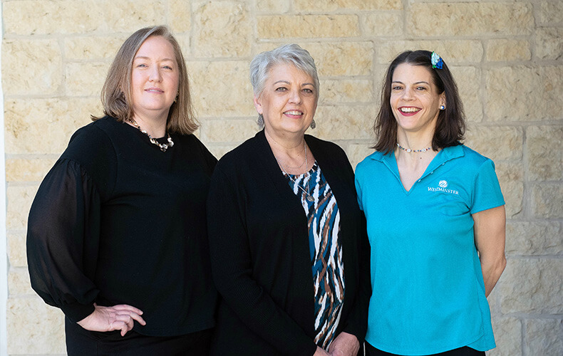 Three staff members standing in front of a stone wall, smiling at the camera.