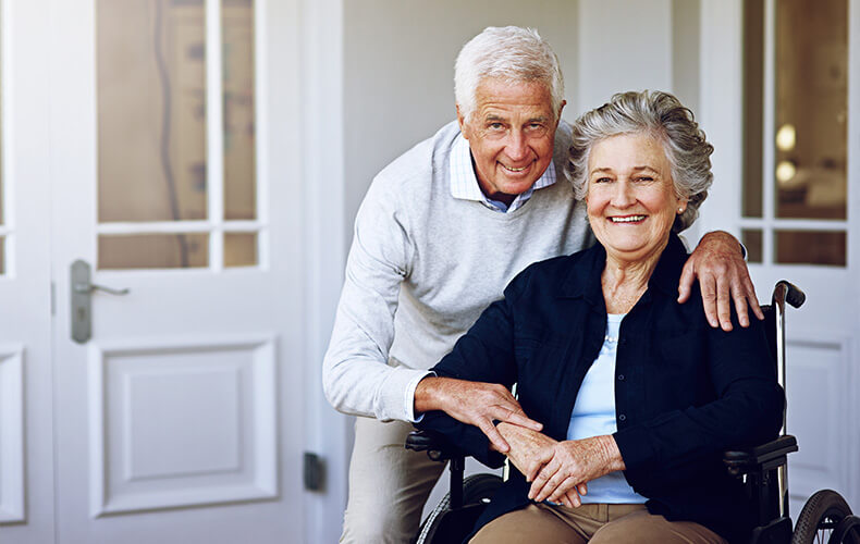 Smiling elderly couple outside a unit, woman in wheelchair, man standing beside her.
