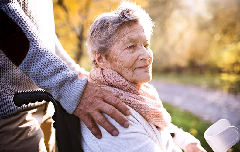 Elderly woman in a wheelchair with a caregivers hand on her shoulder.