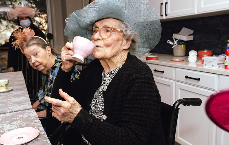 Elderly woman in a blue hat sipping tea in a community dining area.