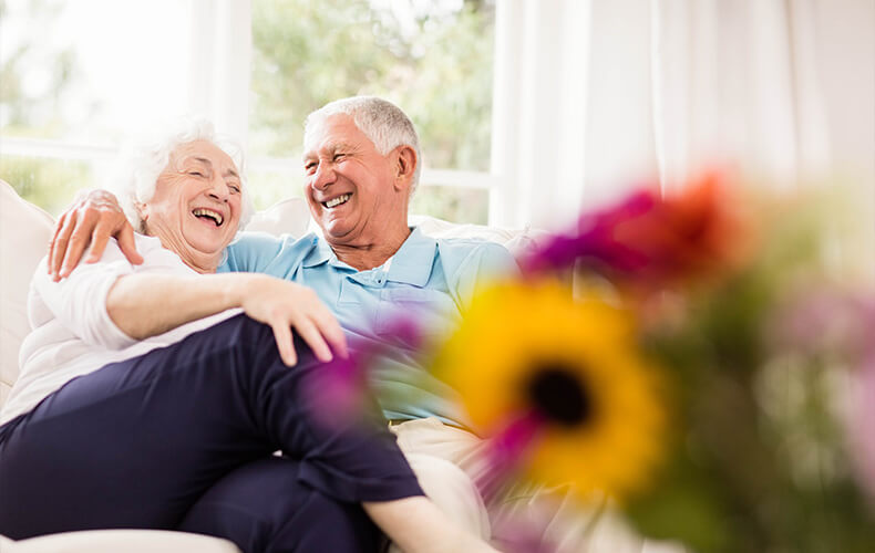 Smiling elderly couple sitting together on a couch with flowers in the foreground.