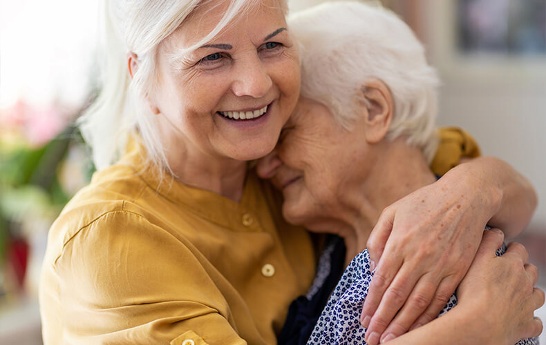 Two elderly women share a warm embrace, smiling joyfully in a cozy setting.