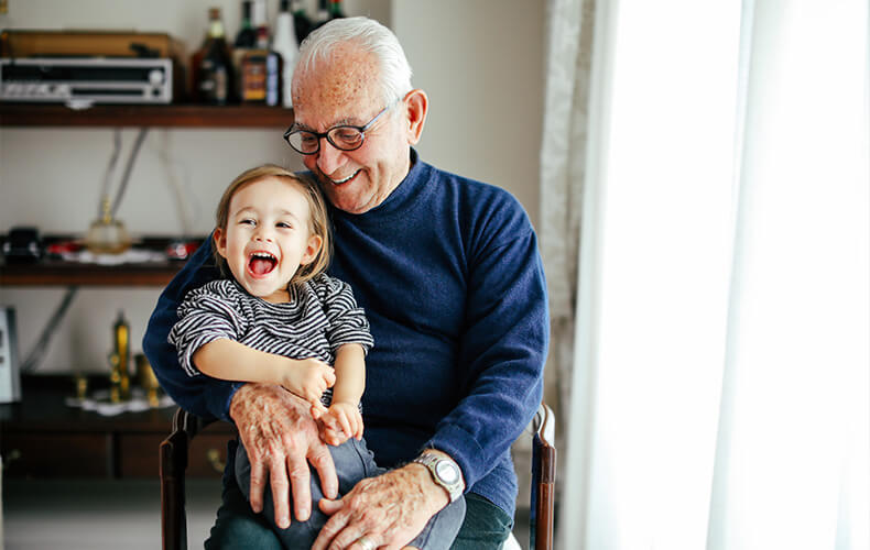 Elderly man joyfully holding a laughing small child inside a well-lit room.
