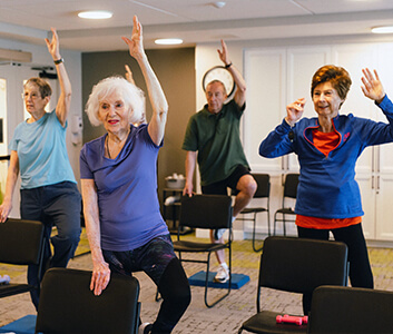 Seniors participating in a lively exercise class, raising arms beside chairs.