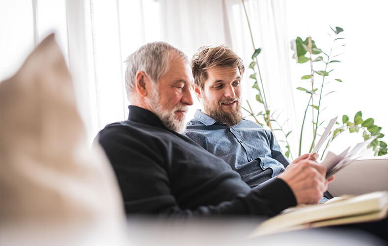 Elderly man and younger man reading documents together on a couch in a bright room.