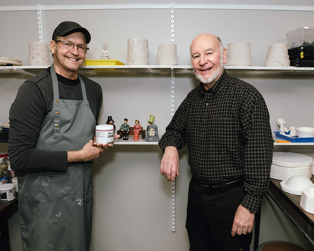Two men smiling in a pottery studio with craft items displayed on shelves.