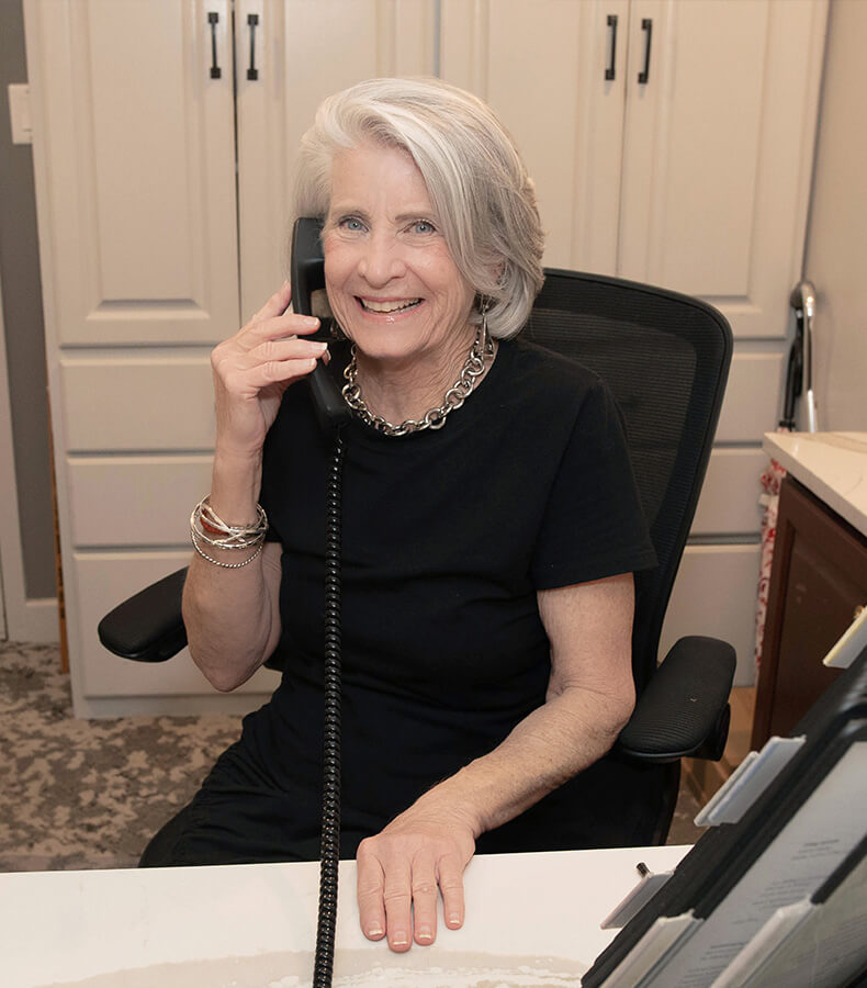 Smiling woman with gray hair on the phone at a reception desk in a communal area.