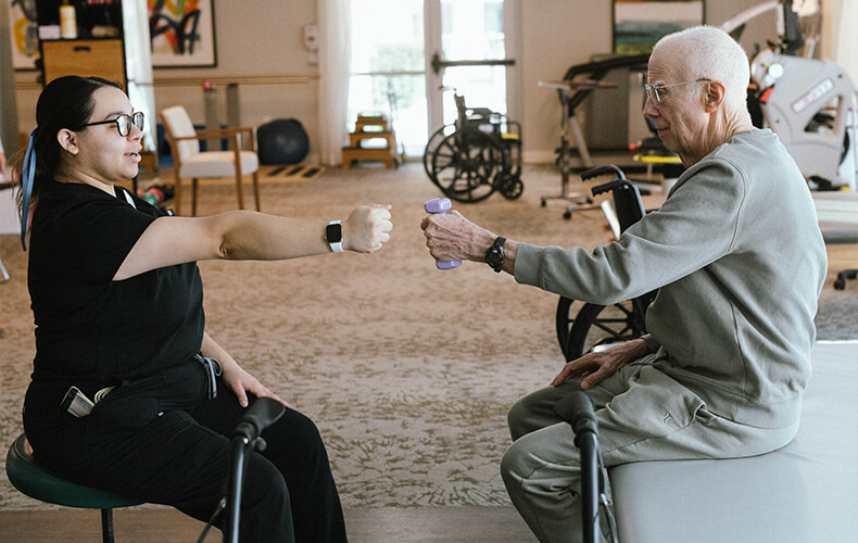 Senior man in physical therapy holding a weight guided by a therapist in a rehab facility.