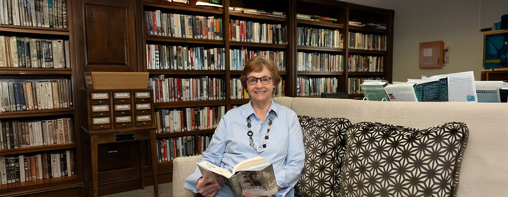Smiling woman in glasses reads a book, sitting on a couch in a library setting.