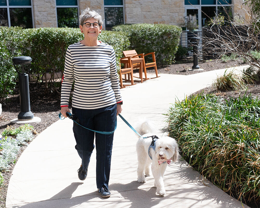 Smiling resident walking a fluffy white dog on a sunny path in the community garden.