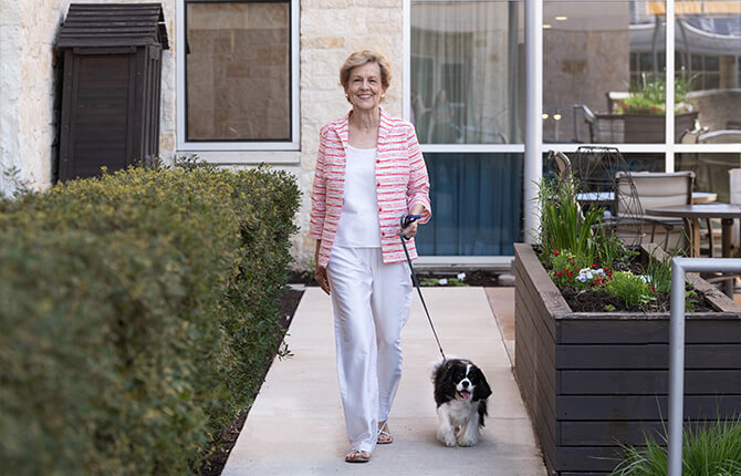 Woman walking a dog on a sunny path beside greenery and modern building units.