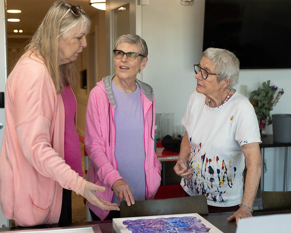 Three older women engaged in conversation while observing art in a community room.