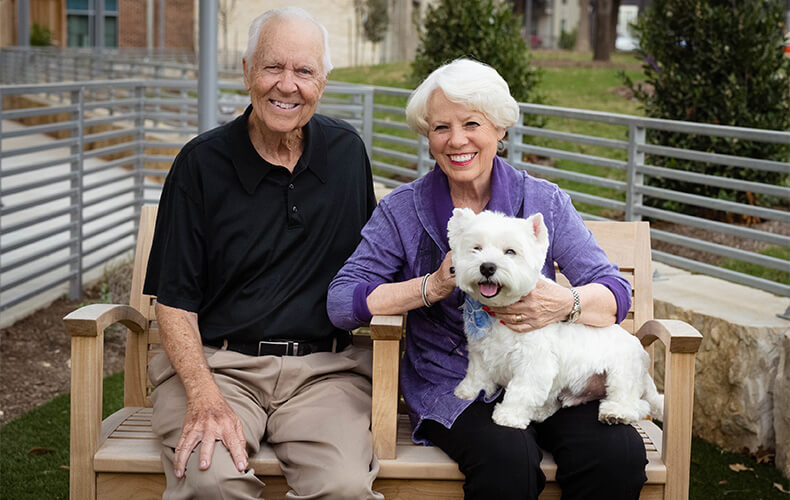 Elderly couple sitting on a bench with a small white dog in a garden area.