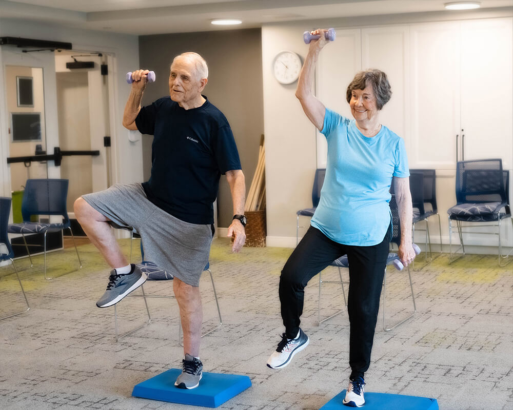 Two seniors exercising indoors with small weights in a modern community center.