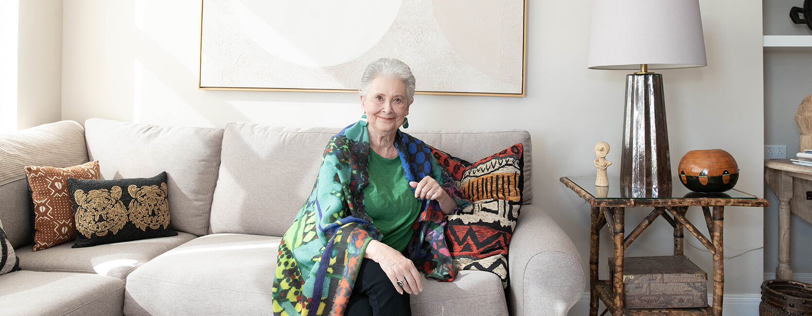 Elderly woman sitting on a sofa in a well-decorated living room with colorful cushions.