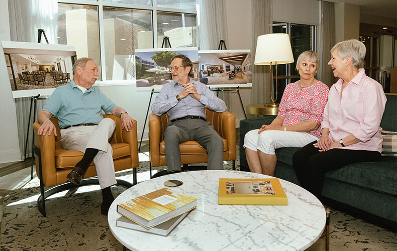 Four seniors discussing community plans in a lounge with architectural photos.