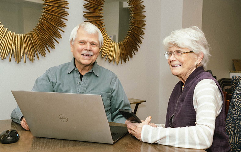 Two seniors smiling while using a laptop and smartphone at a communal table.