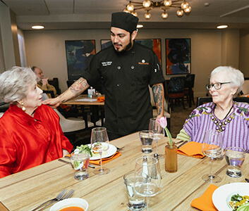 Chef interacting with two residents in a dining area with modern decor.