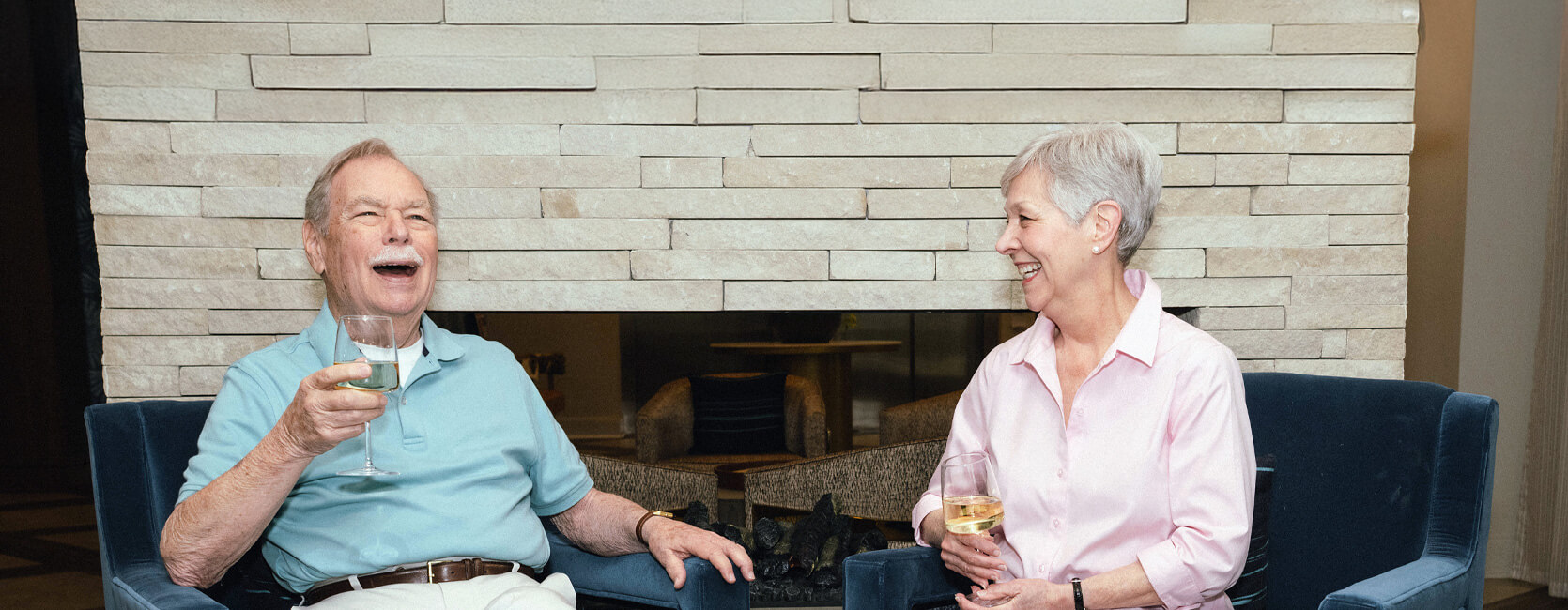 Elderly couple enjoying wine by a cozy stone fireplace.
